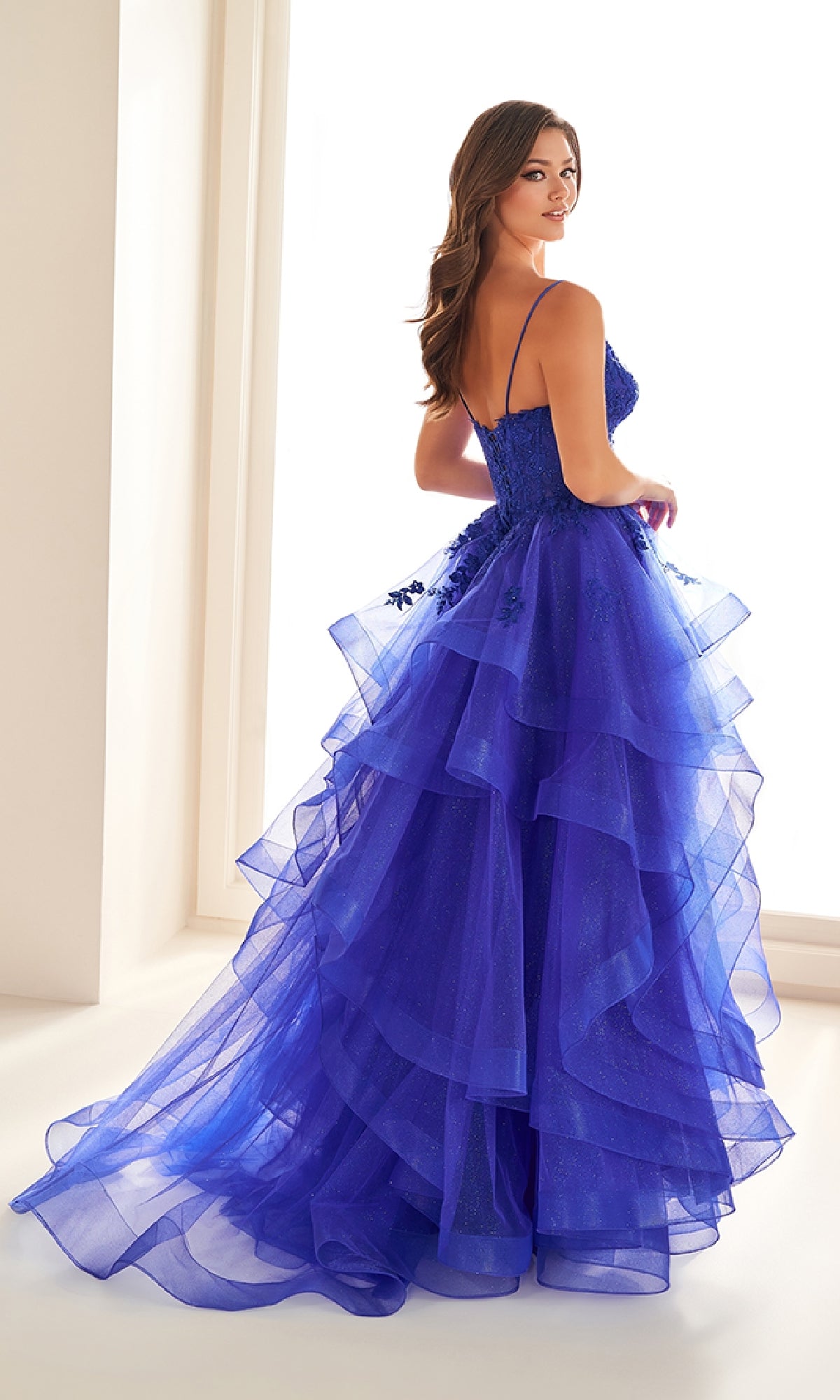 A woman stands in a brightly lit room wearing the Ellie Wilde EW36203 Glitter Tulle Prom Ball Gown—tiered, floor-length, royal blue with floral embellishments, embroidered corset bodice, thin straps, and an adjustable lace-up back.